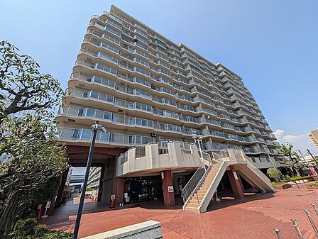 メロディハイム守口の外観は、14階建ての落ち着いた色調のタイル貼りマンションです。青空を背景に、広々としたエントランス前のレンガ敷きの広場と、2階へと続く階段が印象的です。