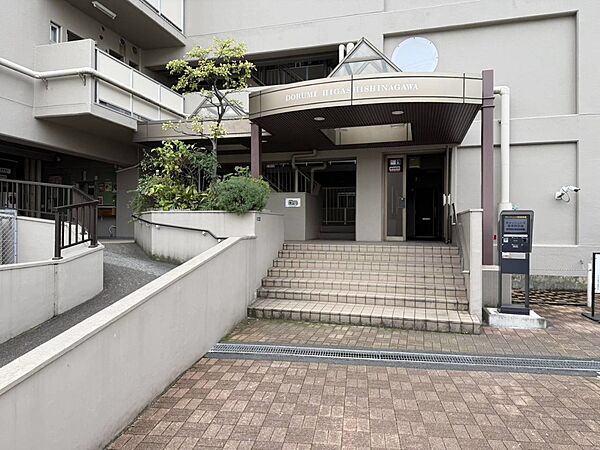 マンション名を掲げた庇と、緑の植栽が迎えるエントランス。落ち着いた外観にタイル敷きのアプローチが調和しています。入口には階段に加えてスロープも設けられ、日々の出入りに配慮された共用部分です。