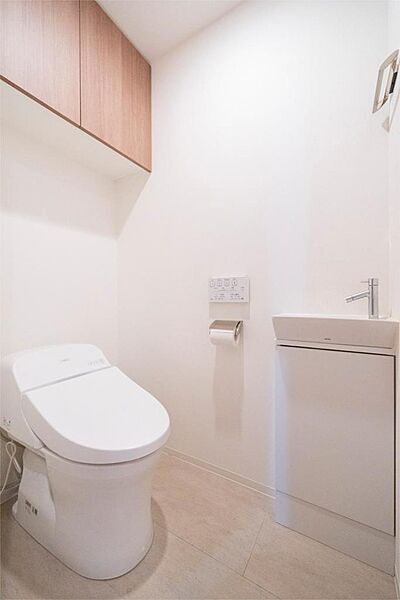 清潔感あふれるホワイトカラーを基調としたトイレ空間。木目調の吊り戸棚が温かみを添え、機能的な手洗い器も備わっています。
