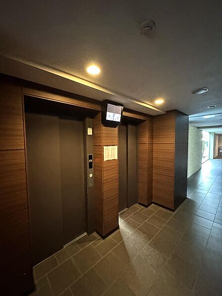 落ち着いた木目調の壁面に包まれたエレベーターホールです。エレベーターは2基設置されており、朝の通勤・通学の時間帯も待ち時間を抑えやすい設計です。防犯カメラの映像を確認できるモニターも備えています。