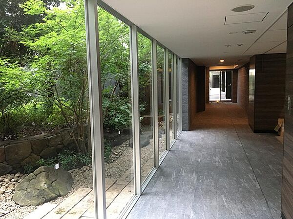 中庭の緑を眺めながら歩ける屋内の共用通路です。ガラス越しに樹々と石組みが広がり、光がやわらかく差し込みます。雨の日もぬれずに移動でき、お子様連れの行き来にも心強いつくりとなっています。