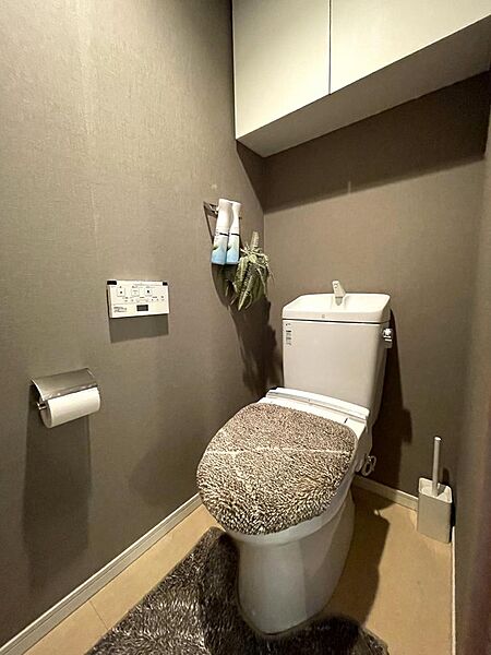 温水洗浄便座付きのトイレです。落ち着いた色合いのアクセントクロスで、くつろげる空間に仕上がっています。上部には吊戸棚が設けられ、トイレットペーパーなどの消耗品もすっきり収納できます。