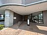 エントランス：円柱と庇で構えたエントランスアプローチ。タイル敷きの通路からガラス扉へ続き、段差を抑えた造りです。両側の開口から自然光が入り、日中も明るさのある共用玄関になっています。