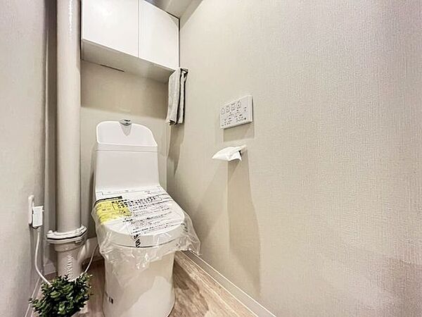 白を基調とした内装に木目調の床材を組み合わせたトイレ空間です。上部には棚が設置されています。手洗い器付きのタンクや、壁掛け式リモコンで操作可能な温水洗浄便座などの設備が備わっています。