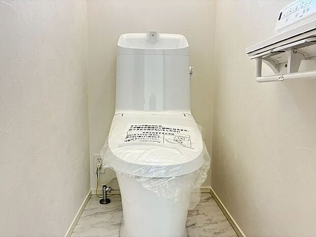白を基調とした清潔感あふれるトイレスペースです。毎日快適にご使用いただける温水洗浄便座を備えています。手洗い一体型のタンクを採用しており、空間を有効活用したすっきりとした印象に仕上がっています。