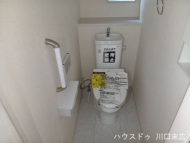 快適な温水洗浄便座を備えた清潔感のある個室。立ち座りをサポートする手すりや、予備のペーパーを保管できる上部収納棚を設けています。