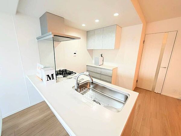 手前に仕切りのないフラットなオープン対面キッチン。背面に食器棚や家電置き場として使えるカップボードと吊戸棚を備えており、調理道具や食器を効率よく収納できる機能的な構造です。