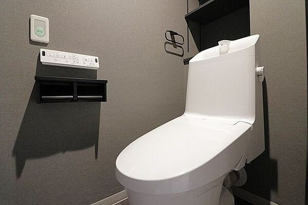 シックなグレーのクロスが落ち着いた雰囲気を演出するトイレ空間。お手入れしやすい手洗い一体型を採用しています。壁掛けリモコンや小物が置ける二連ペーパーホルダーを備えており、毎日の使い勝手も良好です。