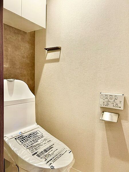 トイレットペーパー等をスッキリ隠せる扉付き吊戸棚を完備！背面のグレージュ調アクセントクロスとウォシュレット一体型便座、スタイリッシュなメタリックパーツで、快適かつ洗練されたレストルームです。