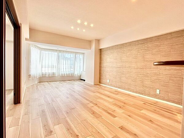 大きな出窓からやわらかな光が差し込む開放的なリビング！質感豊かなアクセントクロスと温もりある木目フロアが洗練された空間を演出します。天井のダウンライトが優しく照らす、くつろぎの居室空間です。