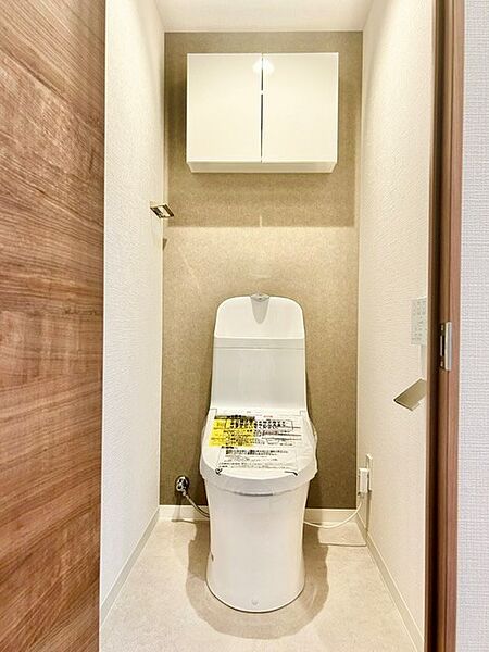 落ち着いたアクセントクロスがおしゃれな温水洗浄便座付きトイレ！上部には吊戸棚を備え、備品をすっきり収納できます。清潔感あふれる空間とスタイリッシュなペーパーホルダーが上質な雰囲気を演出します。