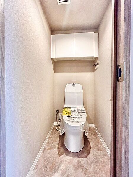 清潔感のある白を基調とした快適なトイレ空間！上部にはトイレットペーパーや掃除用品を隠して収納できる便利な吊戸棚を設置。落ち着いた色合いのクッションフロアが上質な雰囲気を演出する快適な設計です。