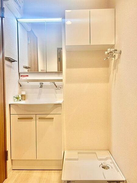 明るい照明が付いた三面鏡付き洗髪洗面化粧台！鏡裏収納や台下のキャビネットにより、生活感を隠してすっきり整理できます。隣には洗濯機置場と便利な吊戸棚を備え、家事動線に優れた清潔感ある空間です。