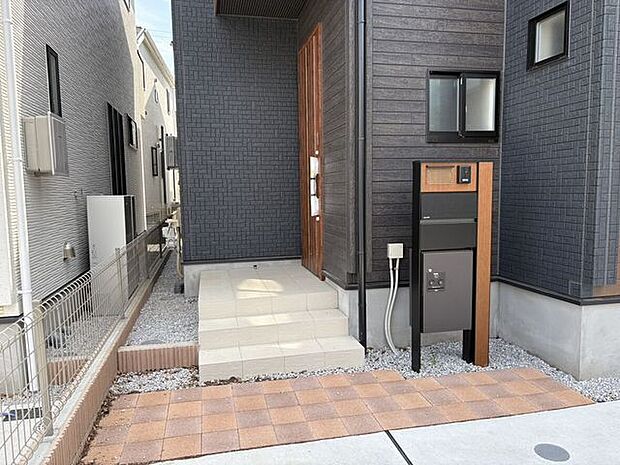 外観と調和した木目調の機能門柱が出迎える、スタイリッシュな玄関アプローチ。宅配ボックスが備わっており、不在時でも安心して荷物を受け取れます。インターホンが奥まった位置にあり、プライバシーにも配慮。