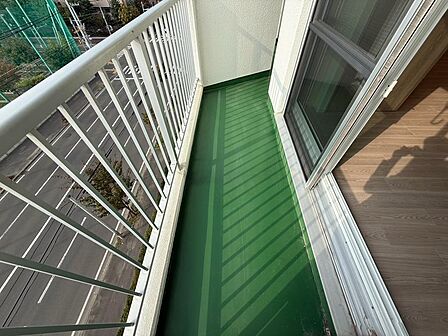 室内から出入りできる、緑色の床が印象的なバルコニー。白色の手すり越しに街並みや道路を望めます。外の空気を感じながら気分転換できる、暮らしに身近な屋外空間。室内とのつながりや奥行きを確認できる写真です。
