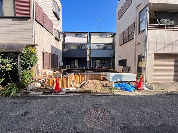 建築中の敷地を正面から確認できる写真。周辺には住宅が建ち並び、落ち着いた街並みの中にあることが分かります。建物の配置や道路との距離感も確認できます。