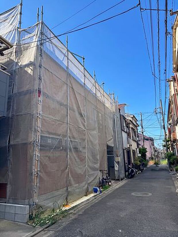 建築中の建物と前面道路を収めた外観写真。建物の大きさや外壁の様子、道路沿いの住宅などが写っており、周辺の街並みも合わせて確認できます。