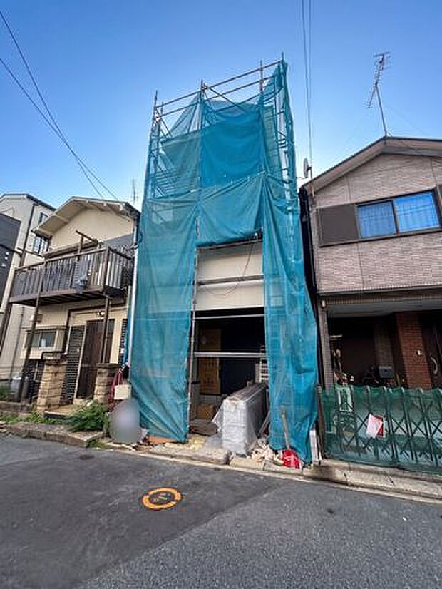建築中の建物を正面から確認できる現地写真。周囲には住宅が建ち並ぶ落ち着いた街並みが広がっており、完成後の外観や周辺環境を想像しながら検討できる一枚です。