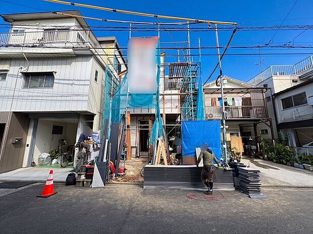 建築中の建物を道路側から見た現地写真。外壁や窓の配置など建物の形状を確認しやすく、周囲の住宅や道路との位置関係も分かりやすい一枚。現地の雰囲気を具体的にイメージできます。