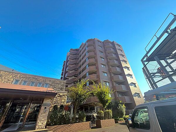 建物正面を見上げる構図で、マンションの高さや外観デザインが分かりやすい写真です。エントランス周辺には植栽も配置されており、緑を取り入れた穏やかな雰囲気が感じられます。
