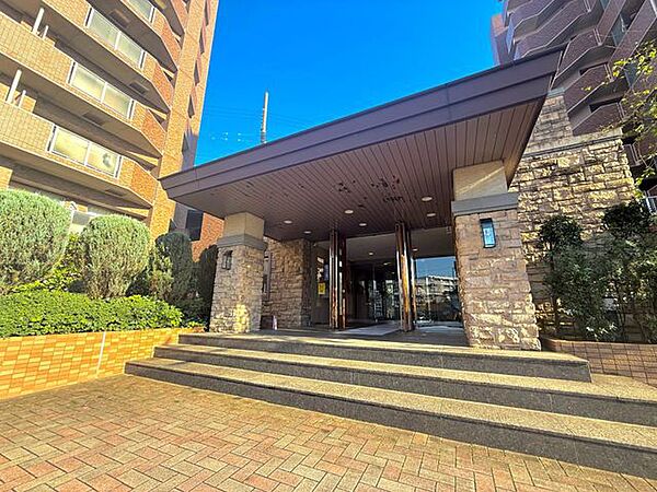 建物のエントランスには屋根付きのアプローチを設けています。石調の外壁や植栽が落ち着いた雰囲気を演出しており、ゆったりとした階段を通って建物内へ入れる造りです。