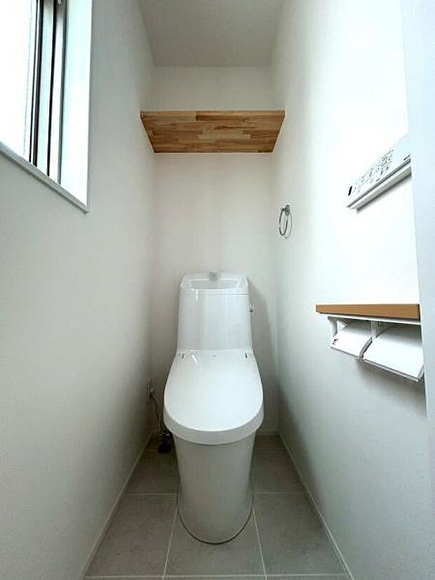 清潔感のある空間が広がるトイレです。窓から自然光が差し込むため明るい印象があります。シンプルなデザインで手入れも行いやすい仕様です。収納棚とペーパーホルダーが備わっており使い勝手にも配慮されています。