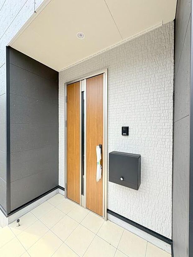 玄関ドアは木目調デザインで外壁とのバランスがすっきりしています。ポストが壁付けで使いやすさが感じられます。タイル張りの玄関アプローチが清潔感を演出しています。屋根付きで雨の日も安心して出入りできます。