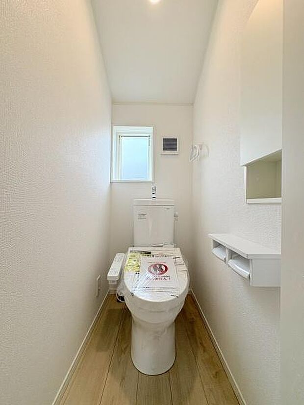 シンプルなデザインで清潔感のあるトイレです。小窓があるため明るさと換気にも配慮されています。床材はナチュラルな風合いが特徴です。ペーパーホルダーや収納スペースも確保されています。