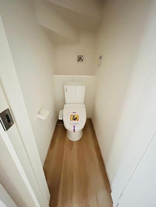 シンプルな空間に設置されたトイレです。清潔感のある内装でお手入れもしやすくなっています。床面には木目調フローリングが採用されています。ペーパーホルダーも備え付けられています。
