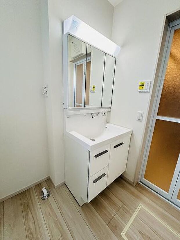 すっきりとしたデザインの洗面台が配置されています。三面鏡が備わっており使い勝手に配慮されています。ホワイトを基調とした清潔感のある空間です。床の木目調が落ち着いた雰囲気を演出しています。