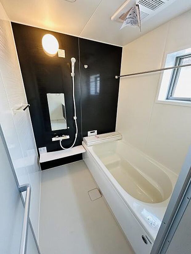 バスルームはシンプルで清潔感があります。黒のアクセントパネルが特徴的です。大きな鏡と十分な広さの浴槽が設置されています。しっかりとした換気設備が備わっています。明るい窓から自然光が差し込みます。
