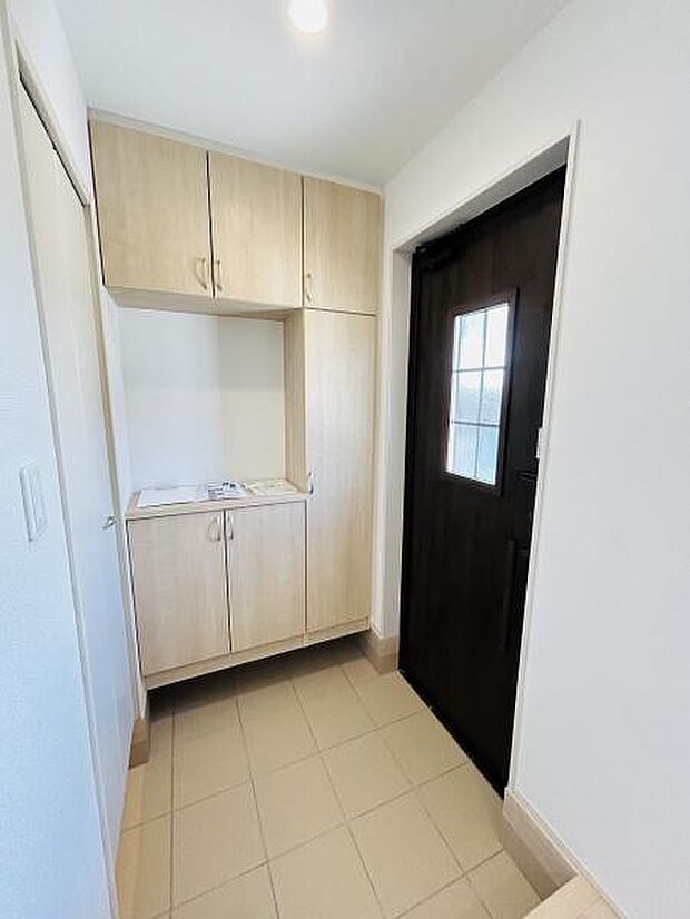 すっきりとした印象の玄関はナチュラルな木目調の収納が備え付けられています。天井まである収納で空間を有効活用できます。明るいタイル床が清潔感を演出しています。ドアの窓から柔らかな光が入ります。