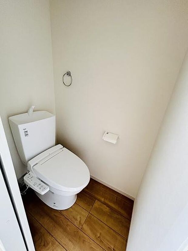 シンプルで清潔感のあるトイレです。壁際に配置されたペーパーホルダーが便利です。フローリング調の床が室内に落ち着きをもたらします。タオルリングも用意されています。ゆとりのある空間設計です。