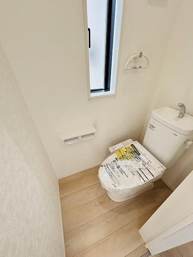 明るい採光のあるトイレです。清潔感のある内装で壁紙や床材もシンプルな仕上がりです。手洗い一体型のタンクが設置されています。紙巻き器やタオルリングも備え付けられています。新しい設備が設置されています。