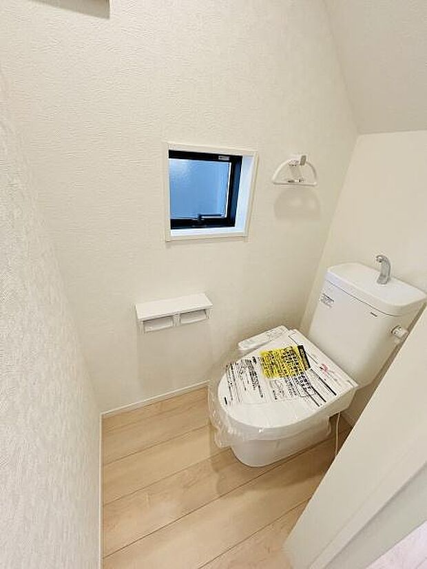 窓が設置されており明るい空間が広がります。壁紙や床材がシンプルなデザインで清潔感があります。ペーパーホルダーやタオルリングなど必要な設備も備わっています。収納や動線にも配慮されたトイレです。