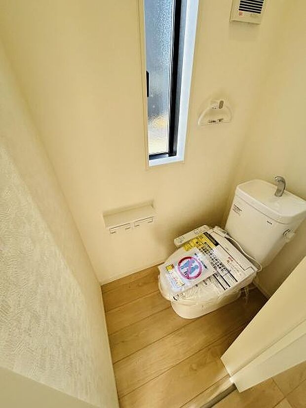 縦長の窓が設けられており自然光が差し込む明るいトイレです。床にはナチュラルな木目調フローリングが使用されています。壁や設備のカラーは統一感があり清潔感のある空間です。