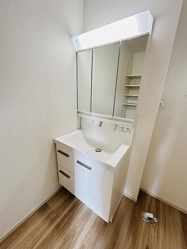 洗面台はホワイトを基調としたシンプルなデザインです。三面鏡が付いていて収納力も魅力的です。床材とのコントラストが空間に落ち着きを与えます。サイドには使い勝手の良い棚も備わっています。