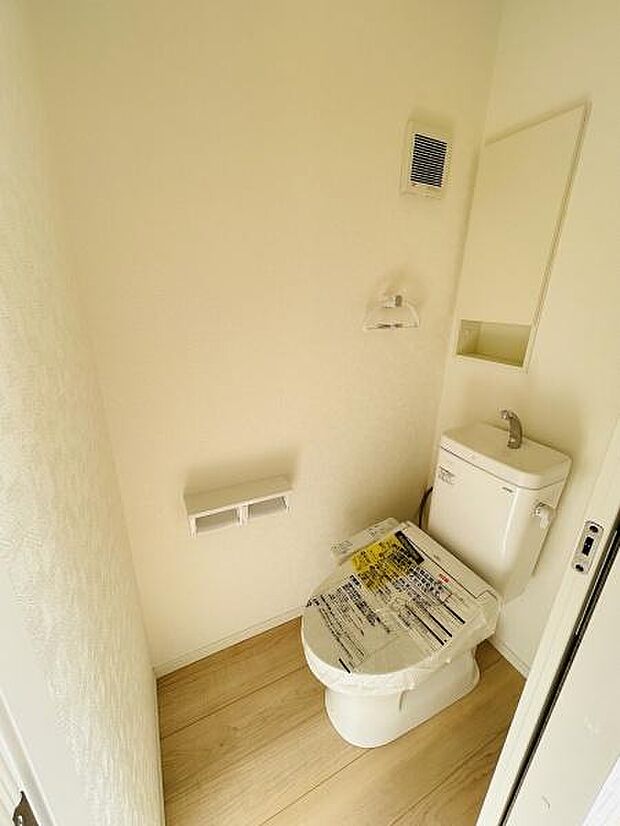 シンプルな内装で清潔感が感じられるトイレです。壁面にはミラーや換気設備も設置されています。ペーパーホルダーや棚の配置が使いやすさを意識した印象です。