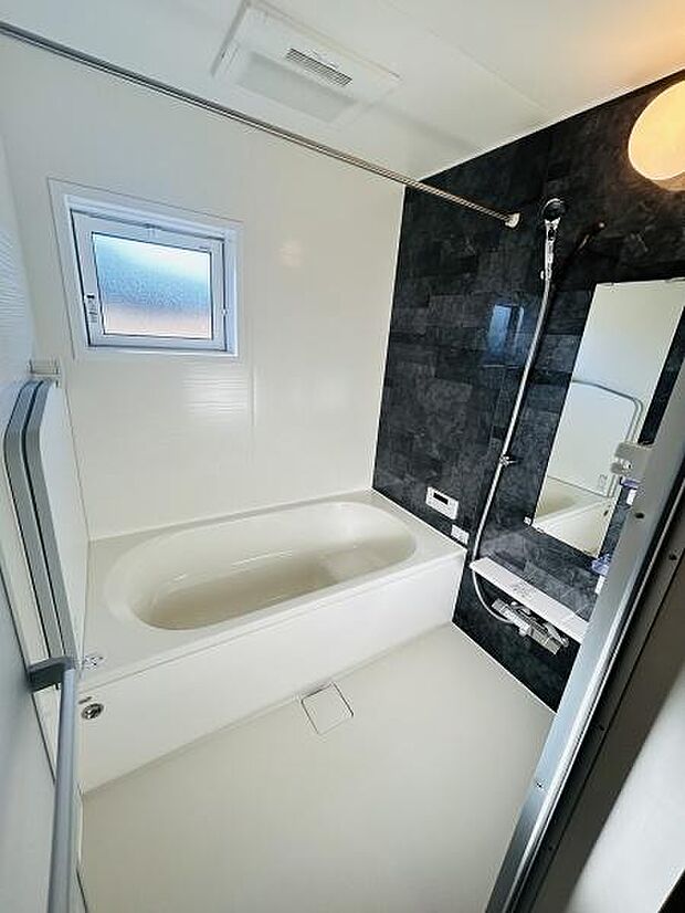 窓が設置されており換気がしやすいバスルームです。ダークカラーの壁面がアクセントとなっています。浴槽スペースがゆったりとした印象です。洗い場と浴槽の動線が確保されています。シャワーや鏡も備わっています。