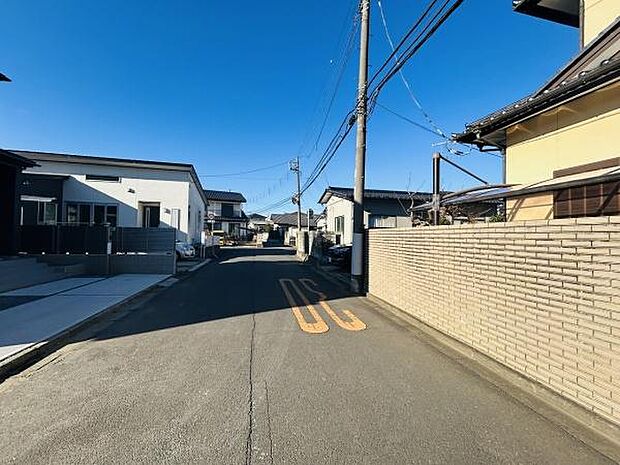 前面道路は整備されておりすっきりとした印象です。周囲には落ち着いた住宅が並んでいます。見通しの良い直線道路が広がっています。電柱や配線も整然と配されています。