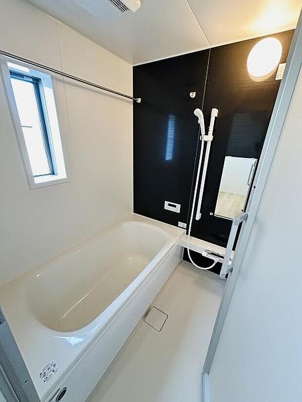 明るい照明と清潔感のある内装が特徴のバスルームです。窓からの自然光も入り換気もしやすい仕様です。黒を基調としたアクセントパネルが空間を引き締めています。浴槽はゆったりとしたサイズ感です。