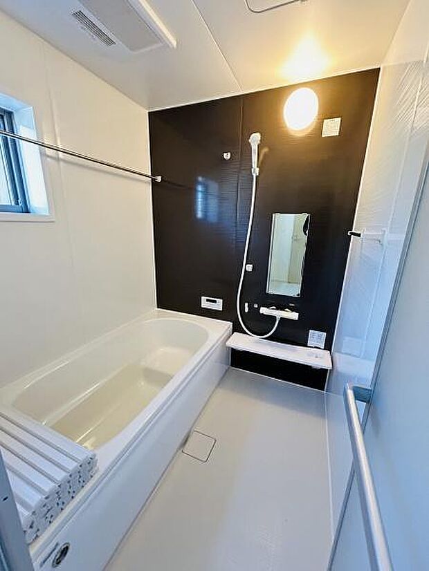 清潔感のあるバスルームです。明るい照明と大きな鏡が特徴的です。落ち着いた色合いのアクセントパネルが印象的です。浴室乾燥用のバーも設置されています。窓があり換気もしやすい空間です。