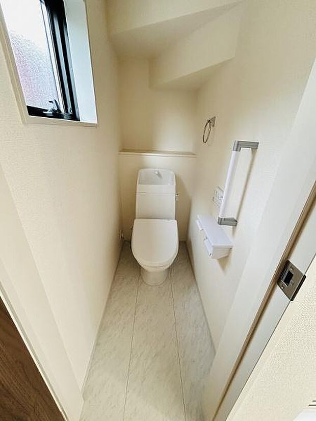 明るい窓付きのトイレで空間がすっきりとまとまっています。手すりも設置されており使用時の安心感があります。床の色合いが清潔感を引き立てています。