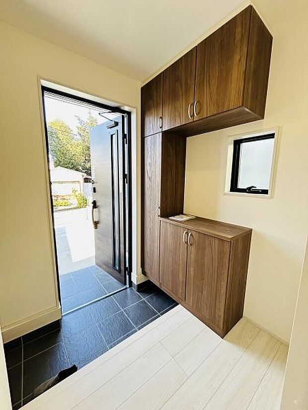 玄関には木目調の収納スペースが設置されておりすっきりとした印象です。ブラックの床タイルがアクセントになっています。明るい空間で窓からも光が入ります。シンプルなデザインが特徴です。