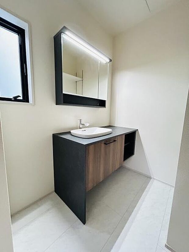 スタイリッシュなデザインの洗面台が印象的です。大きな鏡が空間を広く見せています。ナチュラルな木目調の収納もポイントです。窓があり明るい光が差し込む洗面所です。