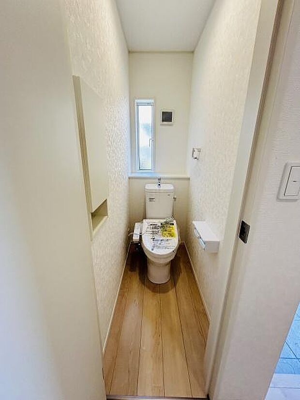 明るい窓付きのトイレで通気性が良く清潔感があります。シンプルな内装が印象的です。壁面収納やペーパーホルダーも備え付けられており使いやすさが考慮されています。床材の色味が空間に温かみをプラスしています。