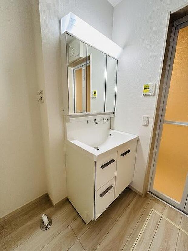シンプルなデザインの洗面台が設置されています。洗面台下の収納が充実しており整理整頓に便利です。三面鏡タイプで使いやすさが感じられます。洗面所の床は落ち着いた色合いとなっています。