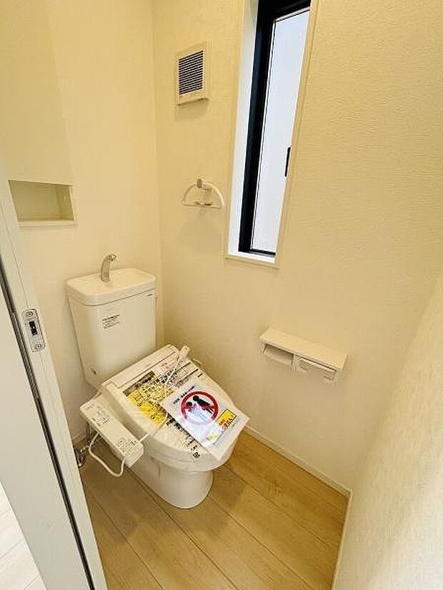 明るい空間が広がるトイレです。木目調の床が優しい印象を与えます。小窓が設けられており換気がしやすい設計です。壁面にはペーパーホルダーや収納スペースも設置されています。