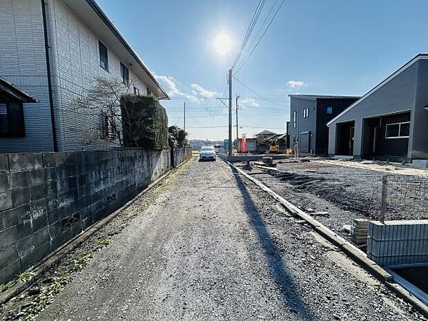 現地の前面道路は幅員が確保されており車の通行も可能です。道路沿いに新築建物や既存住宅が並び落ち着いた雰囲気です。陽当たりの良い道路環境が確認できる写真です。現地の様子がご確認いただけます。