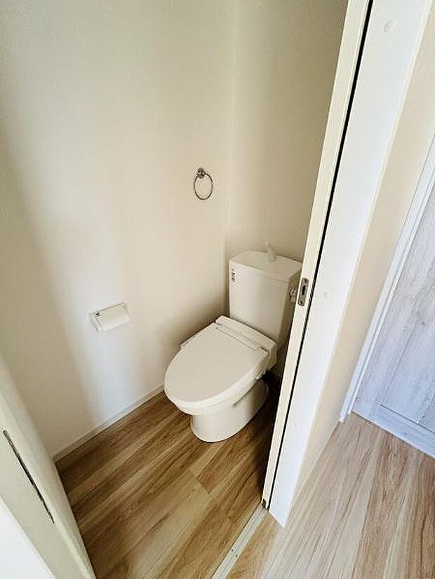 シンプルですっきりとしたトイレ空間です。床の木目調デザインが清潔感を演出しています。ドア付きでプライバシーを確保しやすいレイアウトです。壁にペーパーホルダーとタオルリングが設置されています。